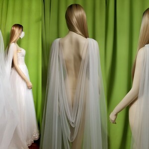 May include: A white tulle bridal cape with a draped back and a long train. The cape is shown on a mannequin against a green background.