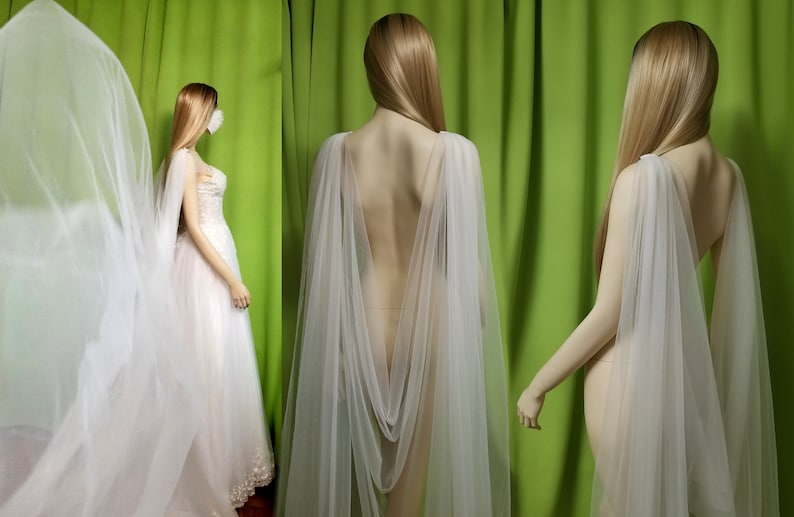 May include: A white tulle bridal cape with a draped back and a long train. The cape is shown on a mannequin against a green background.