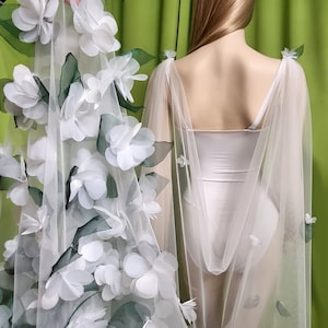 May include: White tulle cape with white flower embellishments and green leaves. The cape has a scoop neck and is sleeveless.