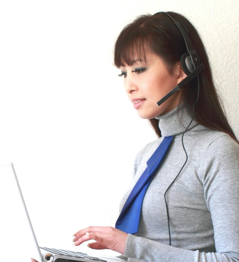 Puede incluir: Una mujer con un jersey gris de cuello alto, una corbata azul y unos auriculares est&aacute; trabajando en un ordenador port&aacute;til.