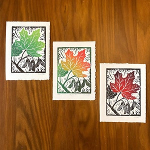 May include: Three woodblock prints of maple leaves in different stages of autumn. The first print shows a green leaf, the second print shows a yellow and orange leaf, and the third print shows a red leaf.