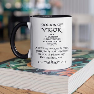 May include: White ceramic mug with a black handle and interior rim. The mug features the text "POTION OF VIGOR" and other text, including "+1 DEXTERITY" and "A BITTER WARMTH fills your belly and ignites in you a flame of DETERMINATION."