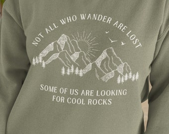 Puede incluir: Una sudadera verde con un gráfico blanco de montañas, un sol y pájaros. El texto dice "Not all who wander are lost. Some of us are looking for cool rocks."