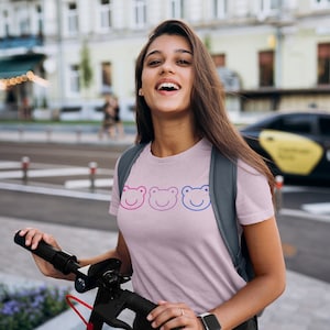 May include: A woman wearing a pink t-shirt with three smiling frog faces in blue, pink, and red. She is holding a black electric scooter and wearing a black backpack and black shorts.