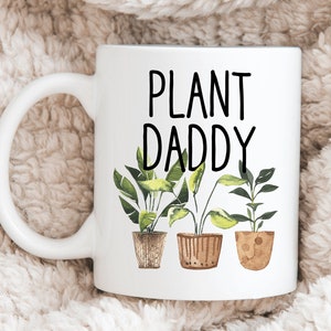 May include: White ceramic mug with a black inscription that reads "PLANT DADDY" and three illustrations of potted plants with green leaves.