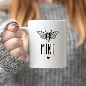 Op de afbeelding: Witte keramische mok met een zwart-witte illustratie van een cicade en het woord "MINE" in zwarte letters met een klein zwart hartje eronder.