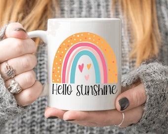 May include: White ceramic mug with a rainbow design and the text "Hello Sunshine". The rainbow is made up of orange, pink, blue, and green stripes with white polka dots. There are three small pink hearts in the center of the rainbow.