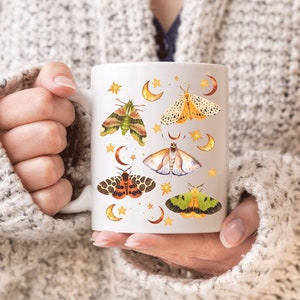 May include: White ceramic mug with a design of colorful moths and crescent moons. The moths are illustrated in a vintage style with intricate details.