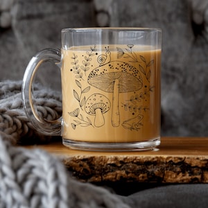 May include: A clear glass mug with a handle, filled with a light brown beverage. The mug has a black and white illustration of mushrooms, leaves, a snail, and a frog.