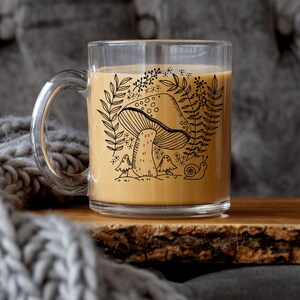 May include: A clear glass mug with a handle, filled with a light brown liquid. The mug features a black line drawing of a mushroom, plants, and a snail. The mug sits on a wooden surface next to a gray knitted blanket.