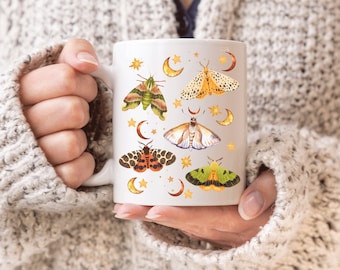May include: White ceramic mug with a design of colorful moths and crescent moons. The moths are illustrated in a vintage style with intricate details.