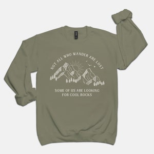 May include: A green long-sleeved sweatshirt with a white graphic of mountains, a sun, and birds. The text "Not all who wander are lost. Some of us are looking for cool rocks." is printed on the sweatshirt.