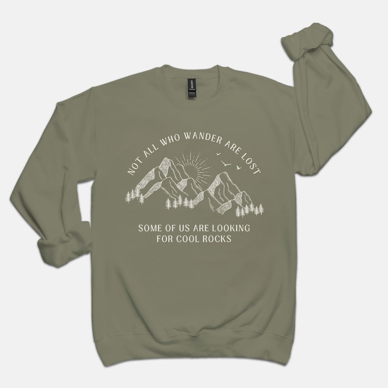 May include: A green long-sleeved sweatshirt with a white graphic of mountains, a sun, and birds. The text "Not all who wander are lost. Some of us are looking for cool rocks." is printed on the sweatshirt.