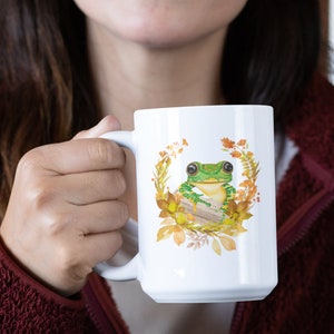 May include: White ceramic mug featuring a green frog perched on a log amidst autumnal leaves and flowers.
