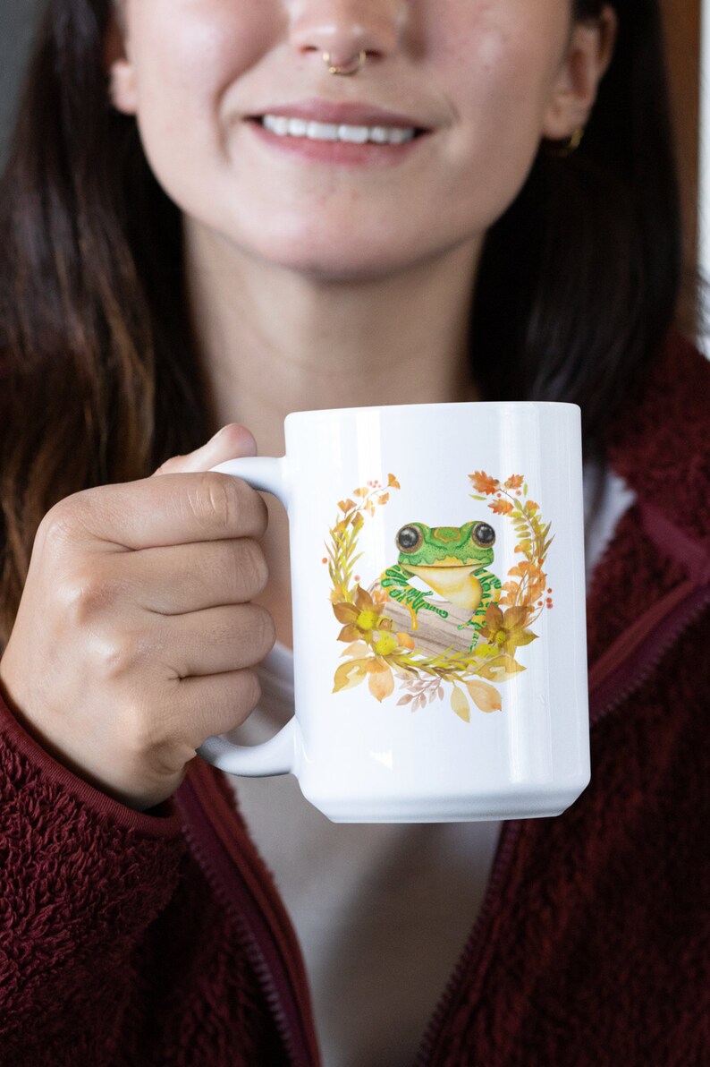 May include: White ceramic mug featuring a green frog perched on a log amidst autumnal leaves and flowers.