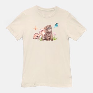 May include: A beige t-shirt with a watercolor illustration of two kittens looking up at a blue butterfly. The kittens are brown and white with pink flowers in the background.