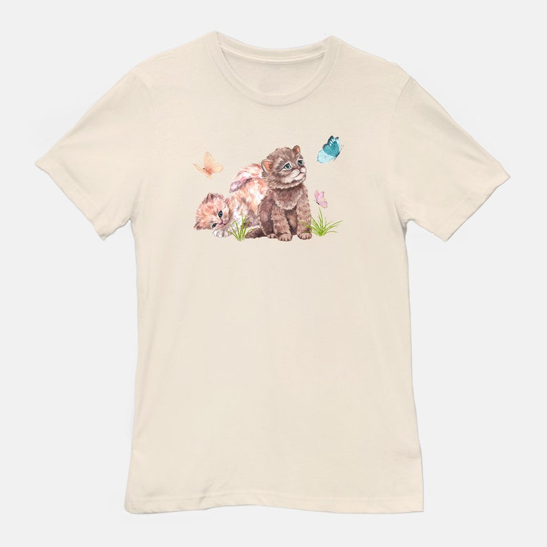 May include: A beige t-shirt with a watercolor illustration of two kittens looking up at a blue butterfly. The kittens are brown and white with pink flowers in the background.