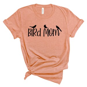 Puede incluir: Una camiseta de color melocot&oacute;n con un gr&aacute;fico negro que dice "Bird Mom" y presenta tres p&aacute;jaros estilizados.