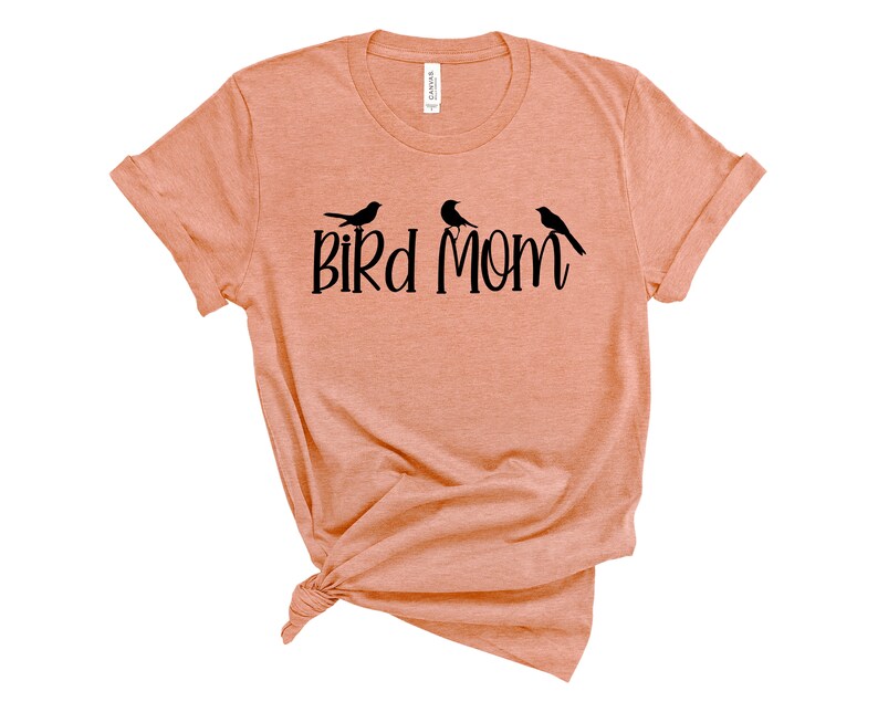 Puede incluir: Una camiseta de color melocot&oacute;n con un gr&aacute;fico negro que dice "Bird Mom" y presenta tres p&aacute;jaros estilizados.