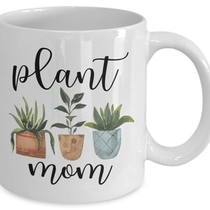 以下が含まれることがあります： 白いセラミック製のコーヒーカップ。黒と白のグラフィックで「plant mom」と書いてあり、緑の葉を持つ鉢植えの植物が3つ描かれています。