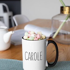 May include: White ceramic mug with a black handle and the name "CAROLE" printed in black on the front. The mug is filled with colorful marshmallows.