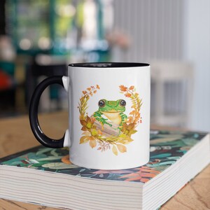 May include: White ceramic mug with a black handle and a black rim. The mug features a green frog sitting on a log surrounded by autumn leaves and flowers.
