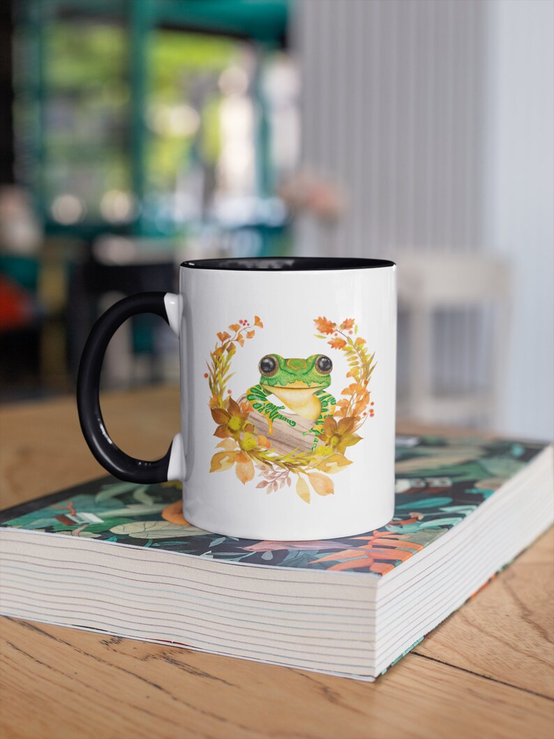 May include: White ceramic mug with a black handle and a black rim. The mug features a green frog sitting on a log surrounded by autumn leaves and flowers.