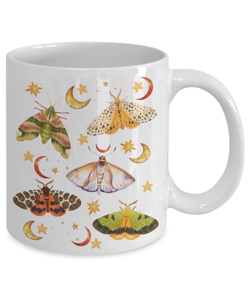 May include: White ceramic mug with a colorful design of moths and stars. The moths are in various shades of green, yellow, orange, and brown. The stars are gold and the background is white.