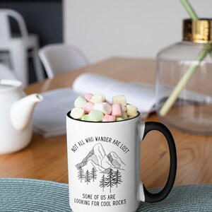 K&ouml;nnte beinhalten: Eine wei&szlig;e Keramiktasse mit schwarzem Henkel. Die Tasse hat einen schwarzen Rand und eine schwarz-wei&szlig;e Grafik von Bergen und B&auml;umen. Der Text auf der Tasse lautet "Not all who wander are lost. Some of us are looking for cool rocks."