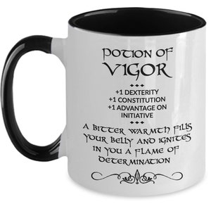 May include: White ceramic mug with a black interior and handle. The mug features the text "POTION OF VIGOR" and other text in a fantasy-themed font. The text includes "+1 DEXTERITY" and "+1 CONSTITUTION".