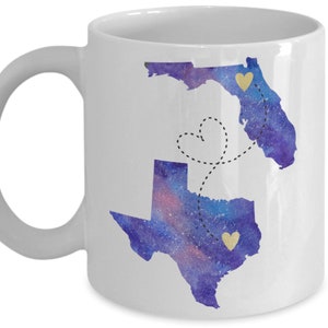May include: White ceramic coffee mug with a watercolor design of the states of Florida and Texas. The states are connected by a dotted line with a heart shape in the middle. The states are filled with a purple and blue watercolor design with gold glitter hearts.