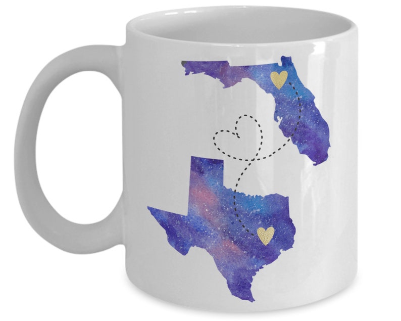 May include: White ceramic coffee mug with a watercolor design of the states of Florida and Texas. The states are connected by a dotted line with a heart shape in the middle. The states are filled with a purple and blue watercolor design with gold glitter hearts.