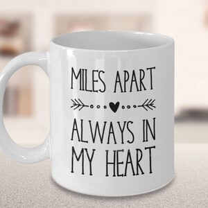 Pu&ograve; includere: Tazza da caff&egrave; in ceramica bianca con testo nero che recita "Miles Apart Always In My Heart" con un piccolo cuore e frecce su entrambi i lati del cuore.
