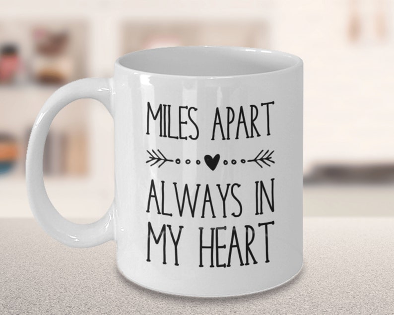 Pu&ograve; includere: Tazza da caff&egrave; in ceramica bianca con testo nero che recita "Miles Apart Always In My Heart" con un piccolo cuore e frecce su entrambi i lati del cuore.