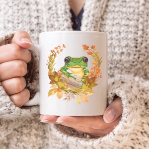 May include: White ceramic mug featuring a green frog perched on a log within a wreath of autumnal leaves and flowers.