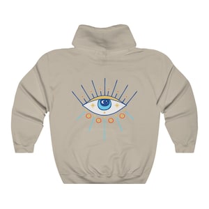 May include: A beige hooded sweatshirt with a blue and yellow evil eye design on the back. The eye has a blue pupil and is surrounded by yellow stars and orange circles.