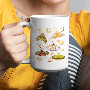 May include: White ceramic mug with a colorful moth and crescent moon design. The mug has a black handle and is filled with a white liquid.