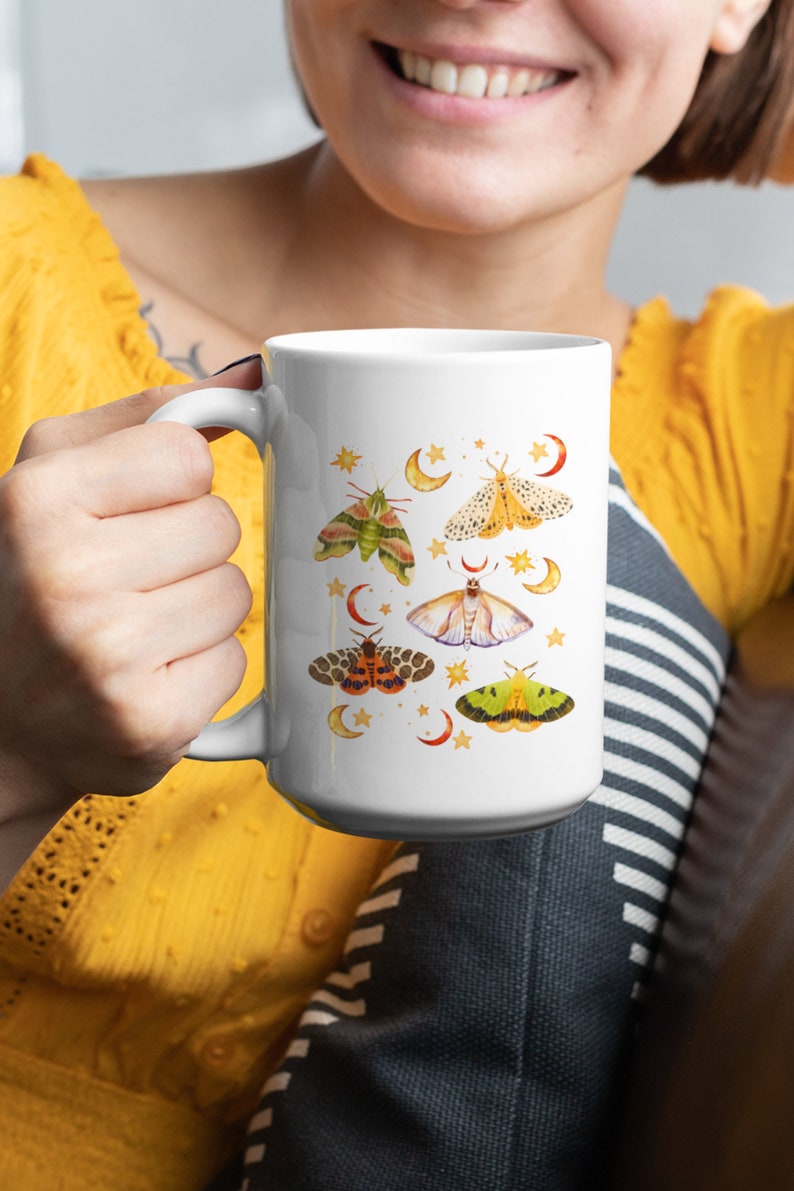May include: White ceramic mug with a colorful moth and crescent moon design. The mug has a black handle and is filled with a white liquid.