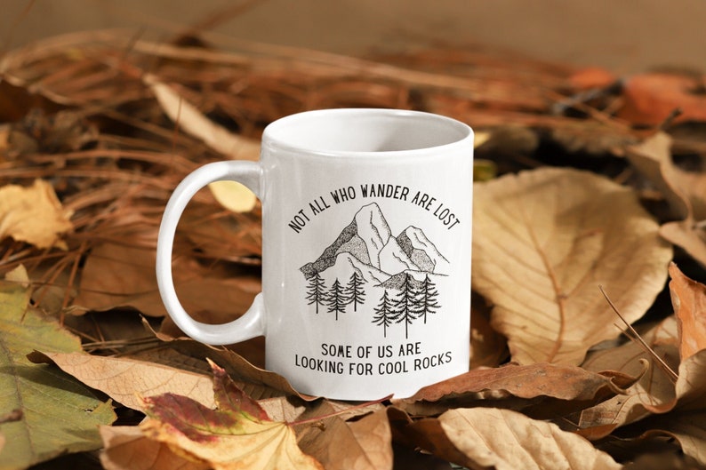 K&ouml;nnte beinhalten: Eine wei&szlig;e Keramiktasse mit einer Schwarzwei&szlig;illustration von Bergen und B&auml;umen. Der Text auf der Tasse lautet "Not all who wander are lost. Some of us are looking for cool rocks."