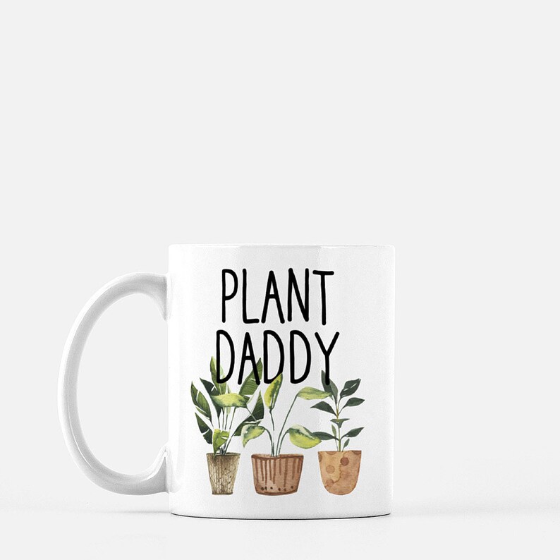 May include: White ceramic mug with a black inscription that reads "Plant Daddy". The mug features a watercolor illustration of three potted plants with green leaves.