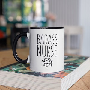 May include: White ceramic mug with a black rim and handle. The mug has the text "BADASS NURSE" in black lettering and a stylized flower design below the text.