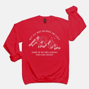 May include: Red long-sleeve sweatshirt with a white graphic of mountains, a sun, and birds. The text reads "Not all who wander are lost. Some of us are looking for cool rocks."