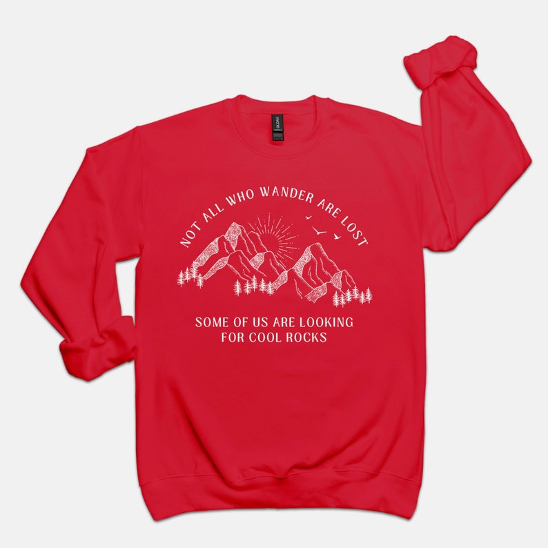 May include: Red long-sleeve sweatshirt with a white graphic of mountains, a sun, and birds. The text reads "Not all who wander are lost. Some of us are looking for cool rocks."