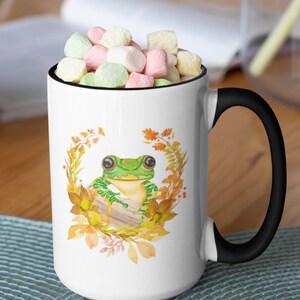 May include: White ceramic mug with a black handle. The mug features a watercolour illustration of a green frog sitting on a log surrounded by autumn leaves.