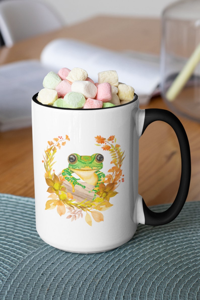 May include: White ceramic mug with a black handle. The mug features a watercolour illustration of a green frog sitting on a log surrounded by autumn leaves.