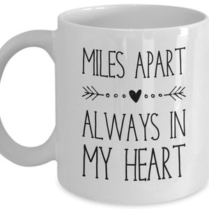 Pu&ograve; includere: Tazza da caff&egrave; in ceramica bianca con testo nero che recita "Miles Apart Always In My Heart" con un piccolo cuore e un design a freccia.