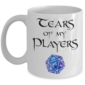 Op de afbeelding: Witte keramische koffiemok met zwarte tekst die "Tears of my Players" zegt en een blauwe en paarse 20-zijdige dobbelsteen.