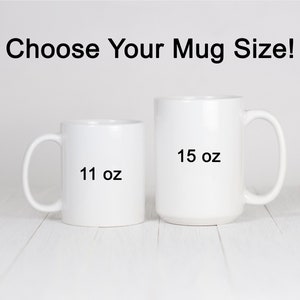 Op de afbeelding: Twee witte keramische mokken, een van 11 oz en een van 15 oz, op een wit houten oppervlak. Kies je mokmaat!