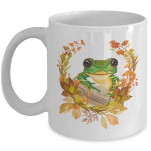 May include: White ceramic mug featuring a green frog perched on a log surrounded by orange and yellow autumn leaves. The frog has large black eyes and a green and yellow striped body.