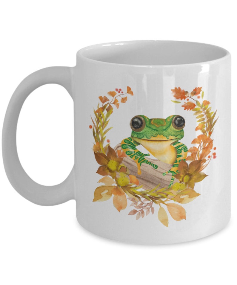 May include: White ceramic mug featuring a green frog perched on a log surrounded by orange and yellow autumn leaves. The frog has large black eyes and a green and yellow striped body.